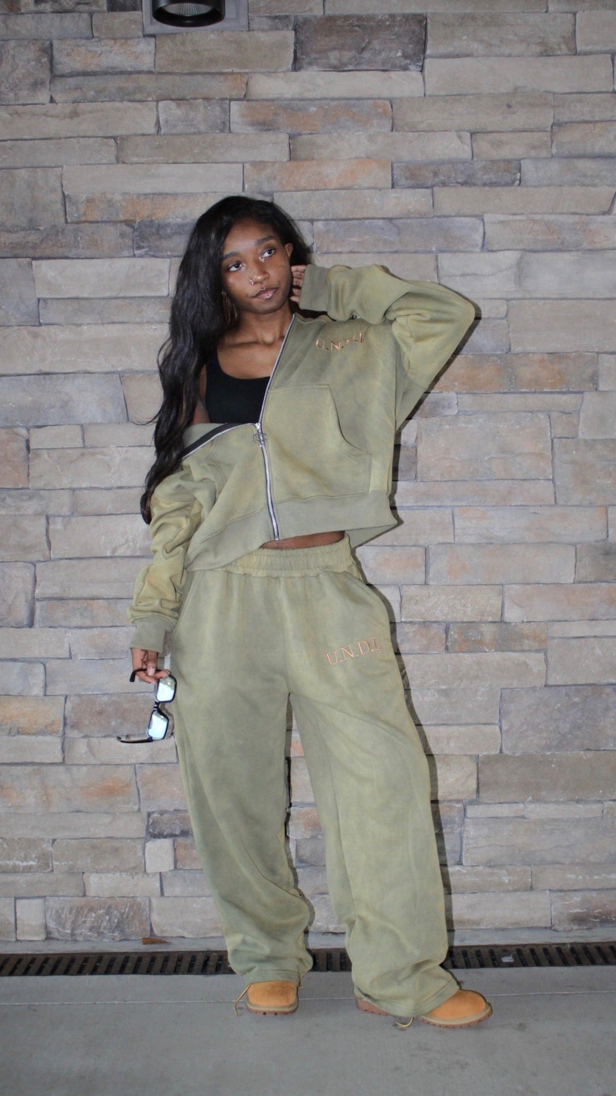 A woman posing in a matching khaki green velour lounge set consisting of a cropped zip-up jacket and wide-leg sweatpants, paired with a black crop top and tan shoes, standing against a stone wall.